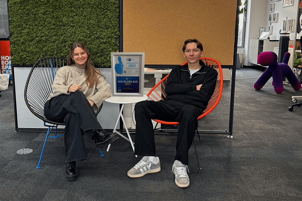 Bettina und Max lächeln im Hauser Showroom. Sie sitzen auf zwei Acapulco Chairs in Orange und Dunkelbraun vor einer Abtrennwand, die aus Kork, Pflanzen, Glas und einem grauen Viereck besteht. Im Hintergrund ist ein Arbeitsplatz und unser Puppy Stuhl zu sehen.