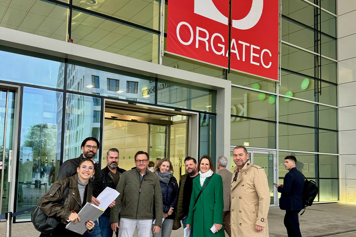 Die Mitarbeiter von Hauser Office Design stehen vor dem Eingang zur Orgatec, einer Messe für Bürodesign und Einrichtungslösungen in Köln. Sie lächeln und posieren unter einem großen roten und weißen Orgatec-Schild, mit moderner Glasarchitektur im Hintergrund.