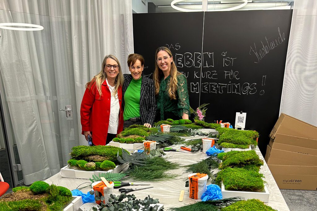 Drei Frauen lächeln vor einer Workshop-Station mit Moosmaterialien, im Hintergrund eine Tafel mit dem Spruch „Das Leben ist zu kurz für langweilige Meetings!“