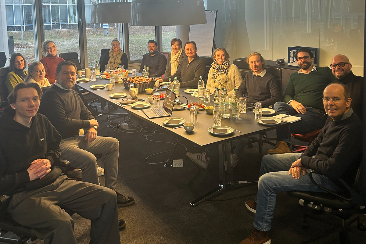 Das Hauser-Team bei einem Meeting in einem modernen Konferenzraum. Die Teilnehmer sitzen an einem großen Tisch, umgeben von großen Glasfenstern, die einen Blick nach draußen ermöglichen. Der Tisch ist mit Getränken, Snacks und Arbeitsmaterialien gedeckt, während die Gruppe in entspannter Atmosphäre zusammenarbeitet