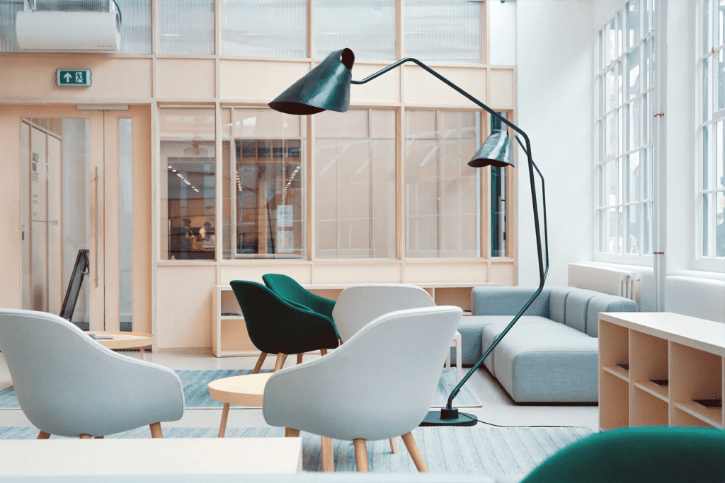 Modernes, offen gestaltetes Büro mit skandinavischem Design. Die Einrichtung umfasst graue und grüne Sessel, eine große Stehlampe mit zwei Lampenschirmen, einen hellblauen Teppich und eine graue Couch. Im Hintergrund sind Glaswände mit Holzrahmen und eine Tür mit Notausgangsschild sichtbar.