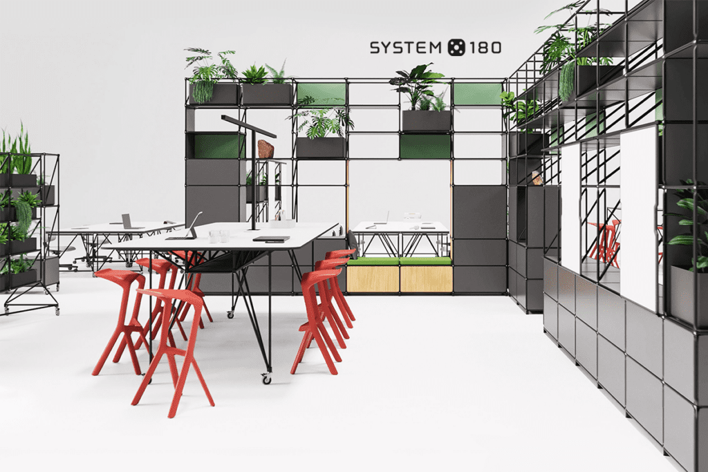 Messestand von System 180 mit Pflanzen und Sitzgelegenheiten – Ein modern gestalteter Raum mit schwarzen Metallrahmen, begrünten Regalen und modularen Büromöbeln. Auffällige rote Stühle setzen Akzente.